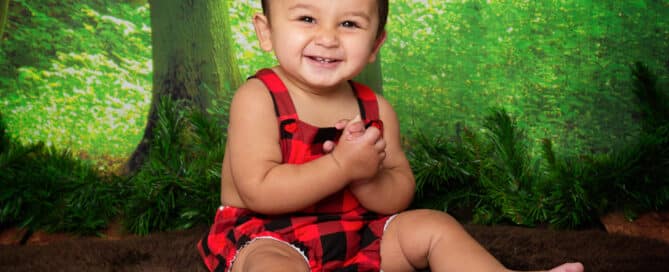 a little boy in a plaid romper sits against a woodland backdrop