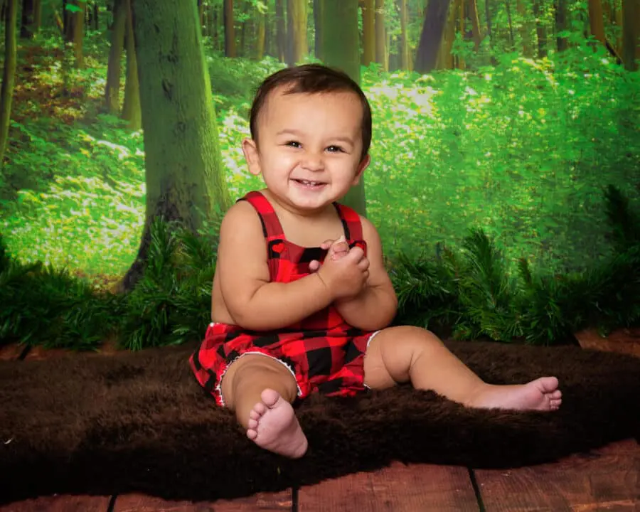 a little boy in a plaid romper sits against a woodland backdrop