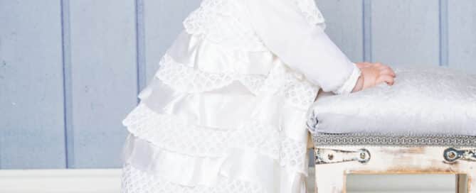 a baby poses in a white christening gown in front of a blue backdrop