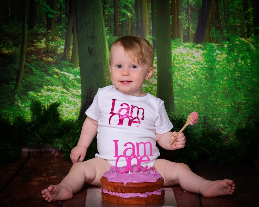a little girl enjoys smashing her cake against a woodland style background at a afirst birthday photo shoot