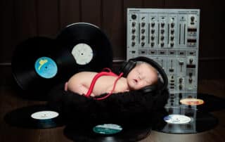 A newborn baby sleeps wearing headphones in front of a mixing deck and surrounded by records
