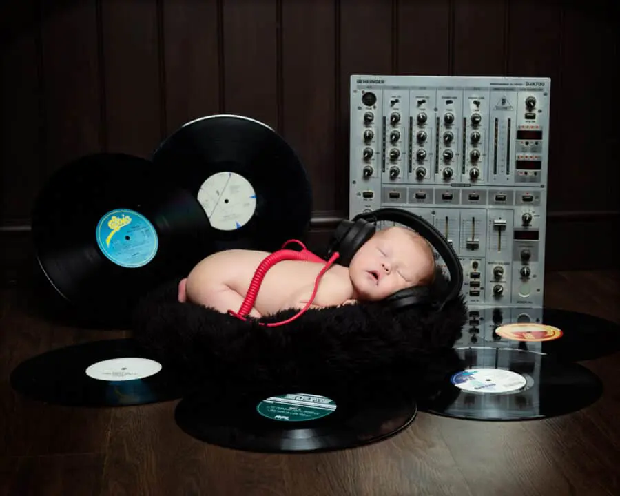 A newborn baby sleeps wearing headphones in front of a mixing deck and surrounded by records