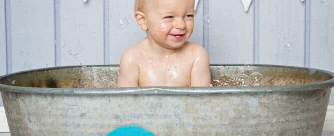 A little boy splashes in a tin tub in front of a blue background and one bunting at a first birthday photo shoot