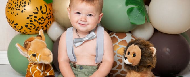 a little boy sits in front of a bundle of jungle themed birthday balloons at a professional photo shoot