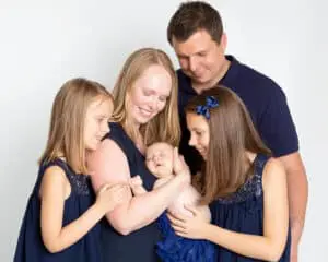 A mum, dad, two older daughters and a newborn arrival pose for a family photo all dressed in navy