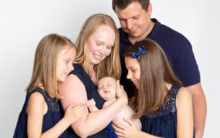 A mum, dad, two older daughters and a newborn arrival pose for a family photo all dressed in navy