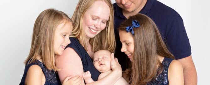 A mum, dad, two older daughters and a newborn arrival pose for a family photo all dressed in navy