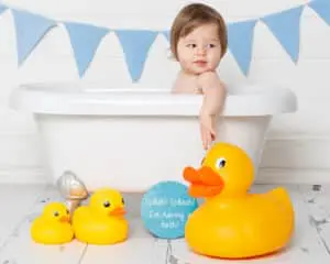 A little boy reaches for a giant yellow rubber duck while he sits in a bathtub at first birthday photo shoot