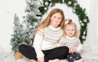 two sisters wear matching jumpers and sit by a giant christmas wreath at a festive photo shoot