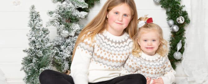 two sisters wear matching jumpers and sit by a giant christmas wreath at a festive photo shoot