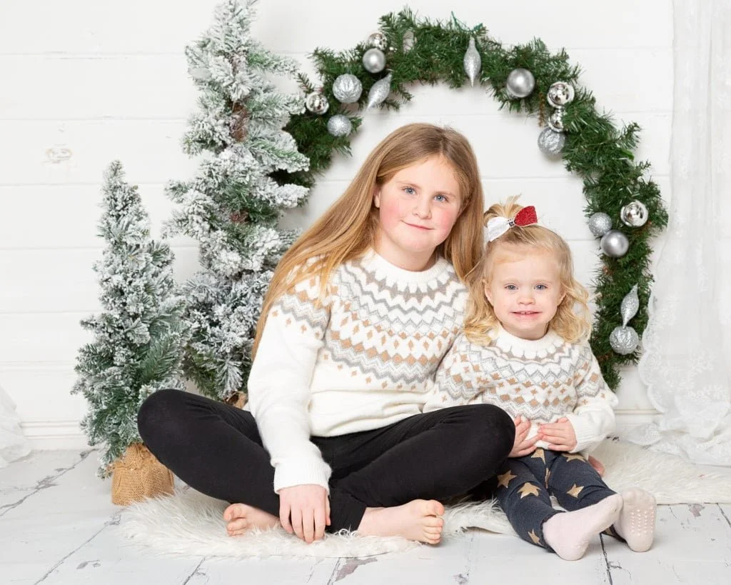 two sisters wear matching jumpers and sit by a giant christmas wreath at a festive photo shoot