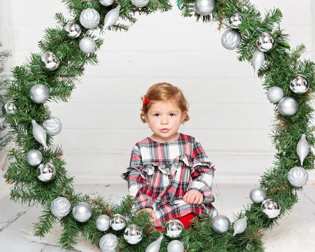 a little girl in a tartan dress sits in the middle of a giant christmas wreath at a christmas photo shoot