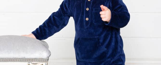 a little boy in a navy romper points at the camera