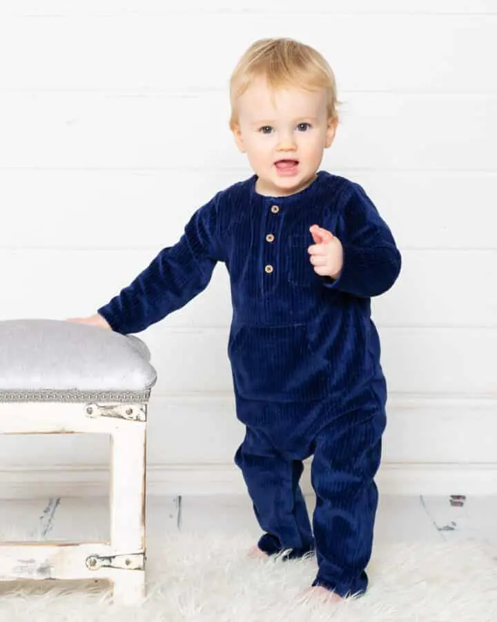 a little boy in a navy romper points at the camera