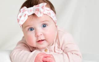 a little girl in a floral pink headband poses on her tummy against a white blanket