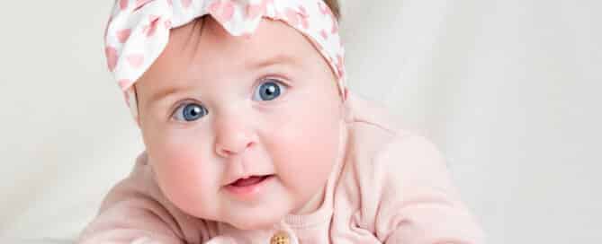 a little girl in a floral pink headband poses on her tummy against a white blanket