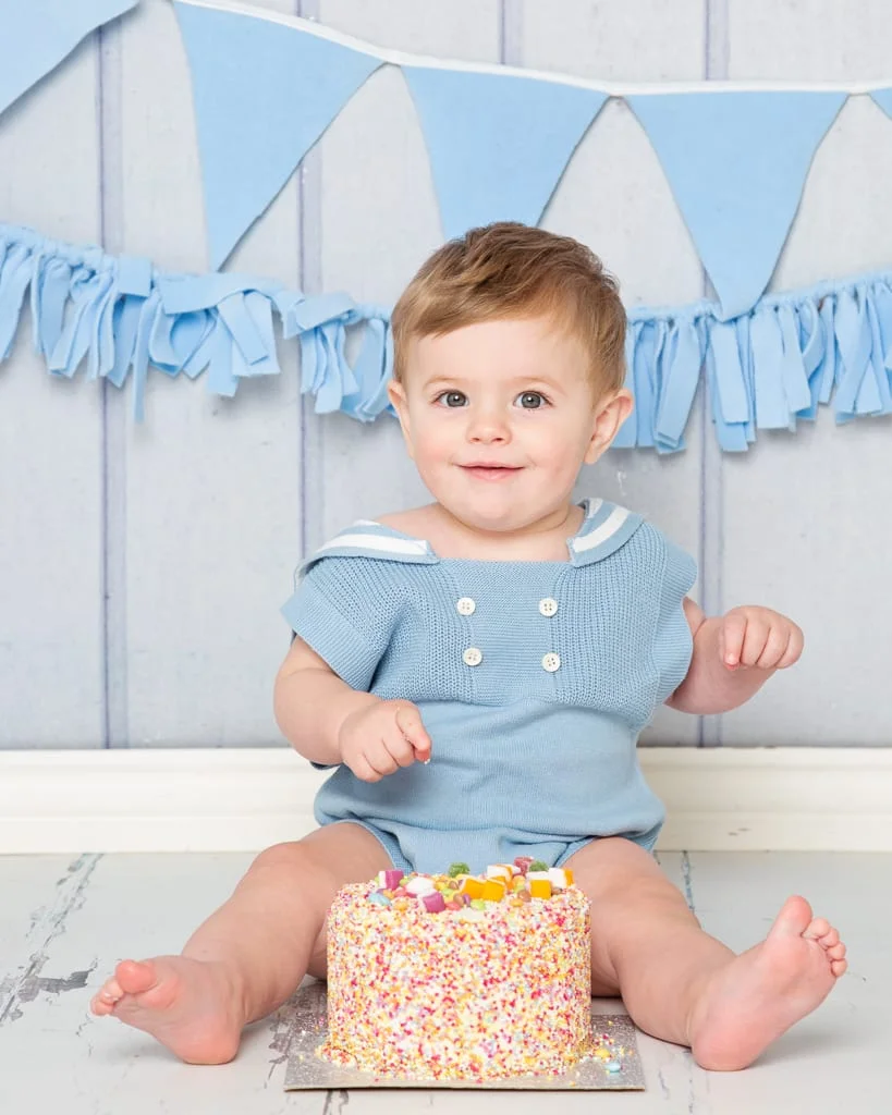 a little boy in a vintage styled blue romper sits by a sprinkle covered cake at his first birthday photo shoot