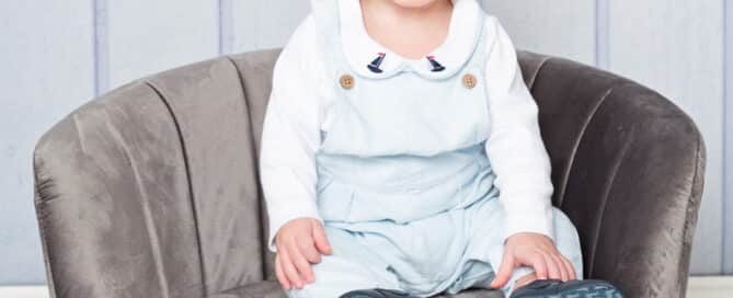 a baby in a blue romper sits on a grey mini chair