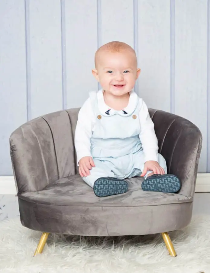 a baby in a blue romper sits on a grey mini chair