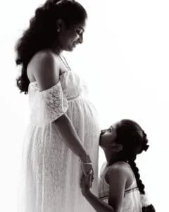 A pregnant lady in a white lace dress hold hands with her daughter and they are looking at each other and smiling