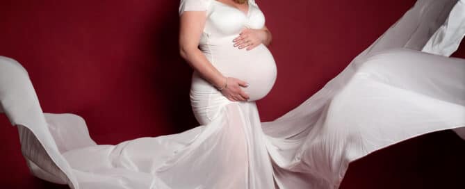 a pregnant woman photographed against a red background in a tight fitting white maternity dress with the train of the dress flying up in the air around her.