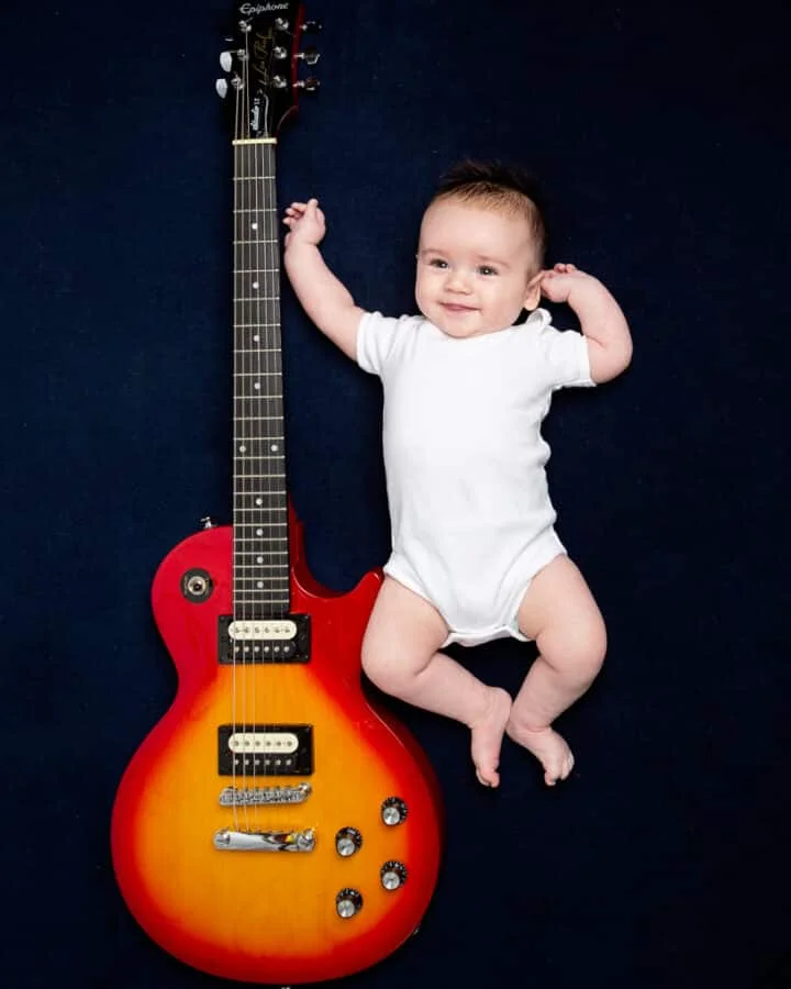 a little boy lies on a jewel toned blanket next to a red guitar
