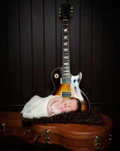 a newborn is partially swaddled and lying on a guitar case with a guitar behind him. He is smiling