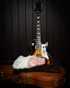 Newborn Photography East Grinstead West Sussex a newborn is partially swaddled and lying on a guitar case with a guitar behind him. He is smiling