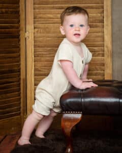 A baby in a cream short romper leans against a wooden footstool at a professional photoshoot against a wooden background