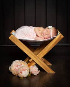 A sleeping newborn is wrapped in a peach swaddle and laying on a wooden hammock with some peach flowers beneath them