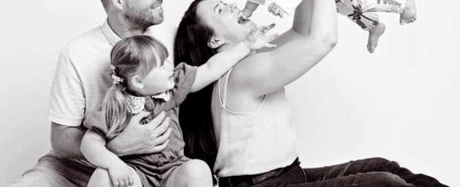 a black and white family portrait where the smallest baby is being flown in the air and everyone is laughing
