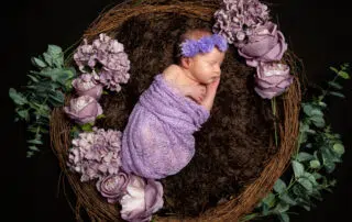 a newborn baby girl wrapped in lilac, wearing a headband and sleeping in a nest prop with lilac flowers