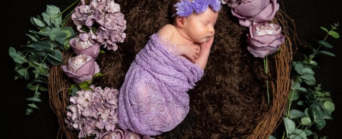 a newborn baby girl wrapped in lilac, wearing a headband and sleeping in a nest prop with lilac flowers