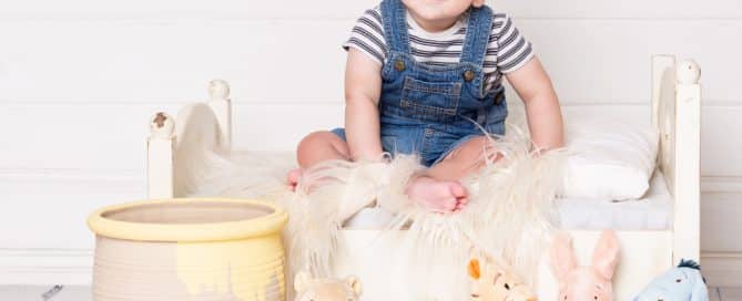 a baby poses on a white bed with a Hunny pot and Winnie the Pooh themed stuffed toys