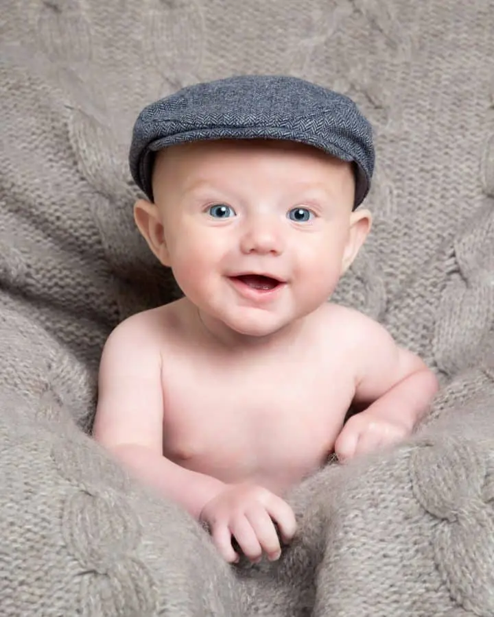 a little boy in a flat cap laughs at the camera against a grey blanket