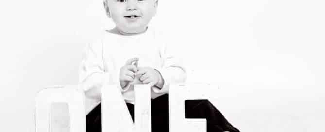 a black and white image of a baby sitting by letters spelling the word ONE