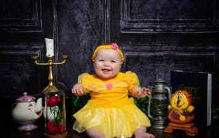 a baby smiles in a Belle dress with Beauty and the Beast Props