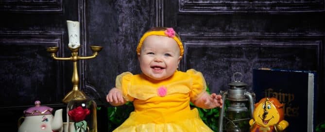 a baby smiles in a Belle dress with Beauty and the Beast Props