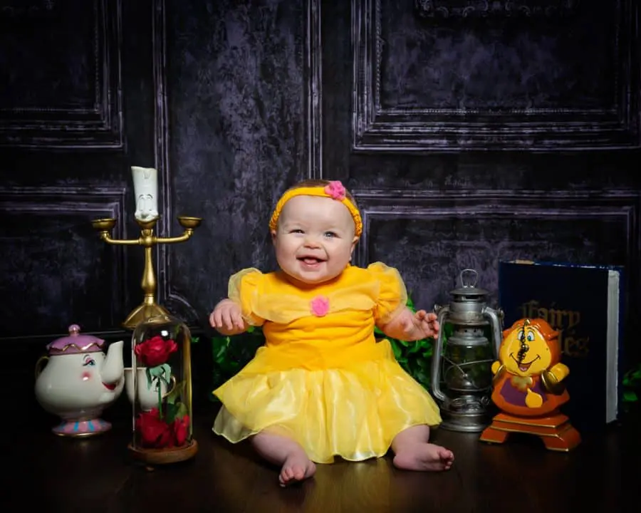 a baby smiles in a Belle dress with Beauty and the Beast Props