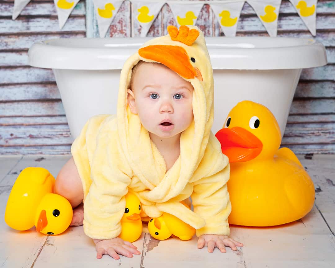 A baby in a duck dressing gown posing with some ducks in front of a miniature bath