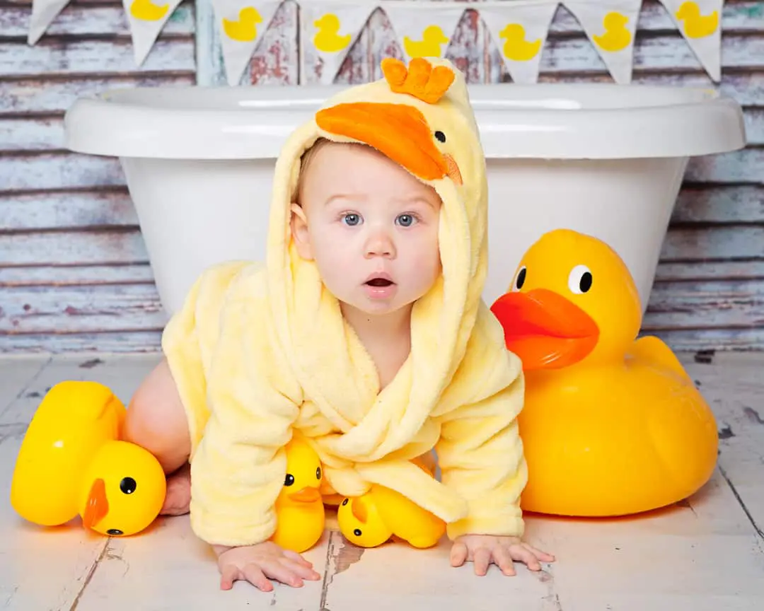 A baby in a duck dressing gown posing with some ducks in front of a miniature bath