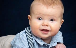 a little boy in a shirt and braces poses on his tummy with a dark background