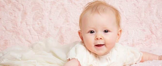 a little girl in a christening dress against a pink blanket backdrop