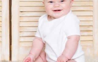 a baby in a classic white vest against some light wood shutters