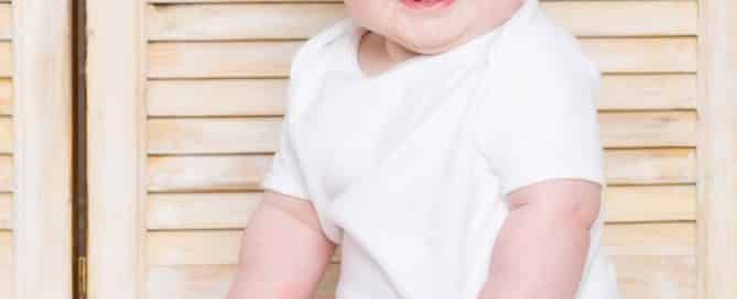 a baby in a classic white vest against some light wood shutters