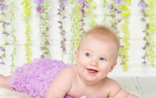 A young baby lying on their tummy in a purple pettiromper in front of a flower wall