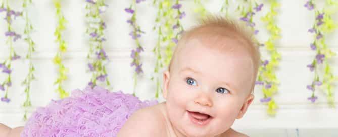 A young baby lying on their tummy in a purple pettiromper in front of a flower wall
