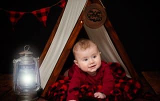 a baby in a miniature camping tent prop set against a black background
