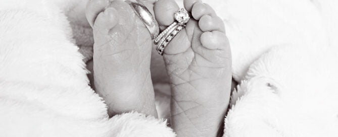 a black and white image of newborn feet with parents wedding rings wrapped in a white blanket