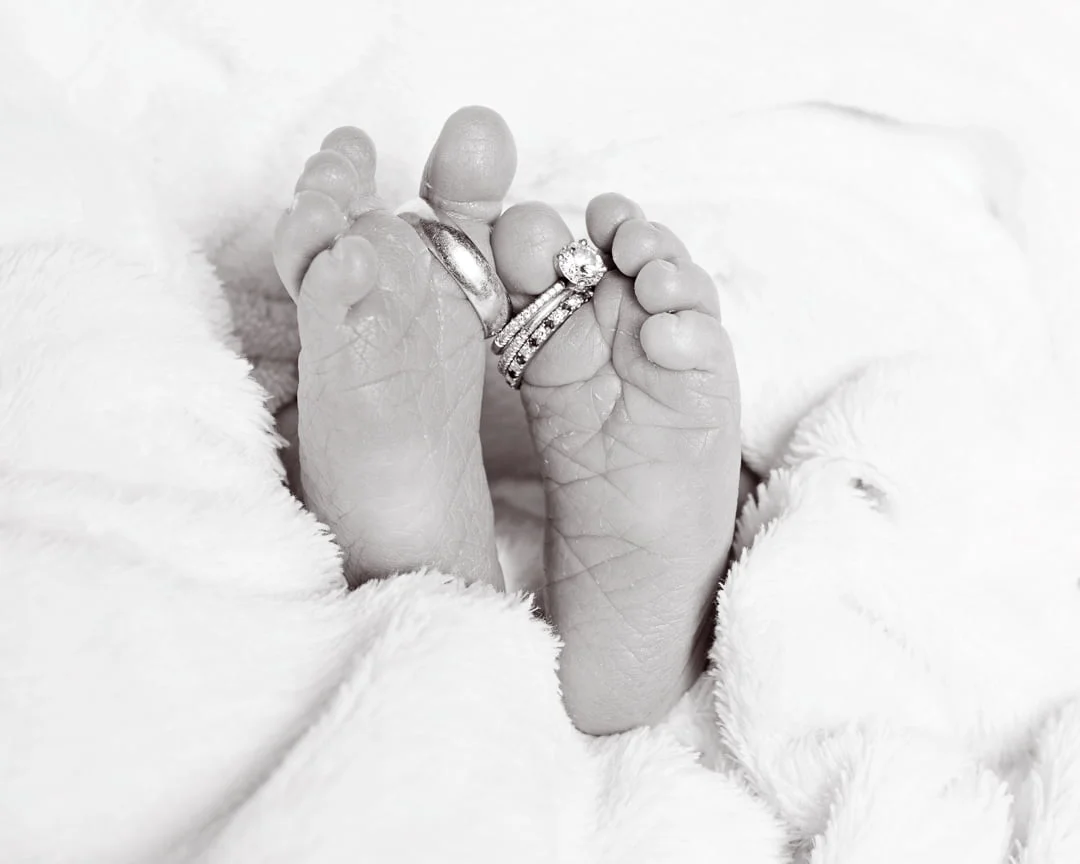 a black and white image of newborn feet with parents wedding rings wrapped in a white blanket